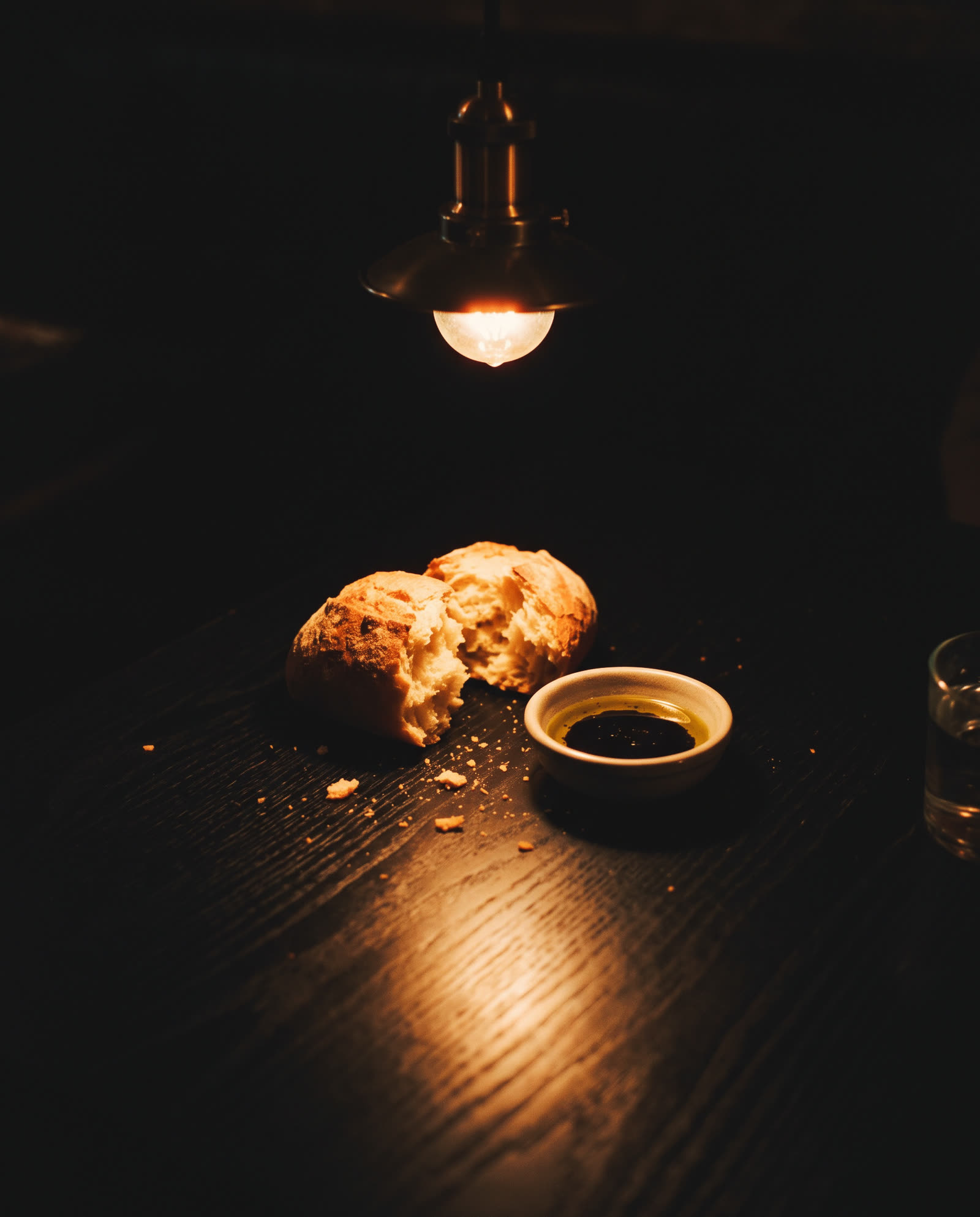 Torn bread and a dish of olive oil on a black table in one pool of lamp light