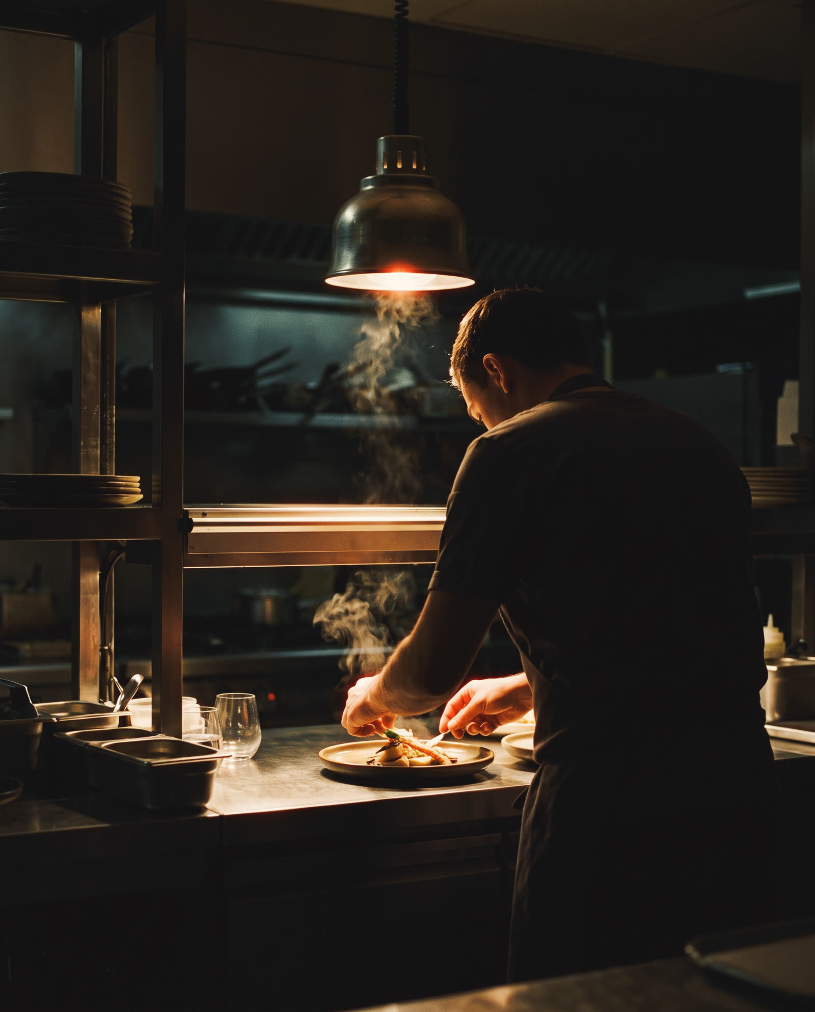 A cook seen from behind at the pass, plating under one lamp, steam rising