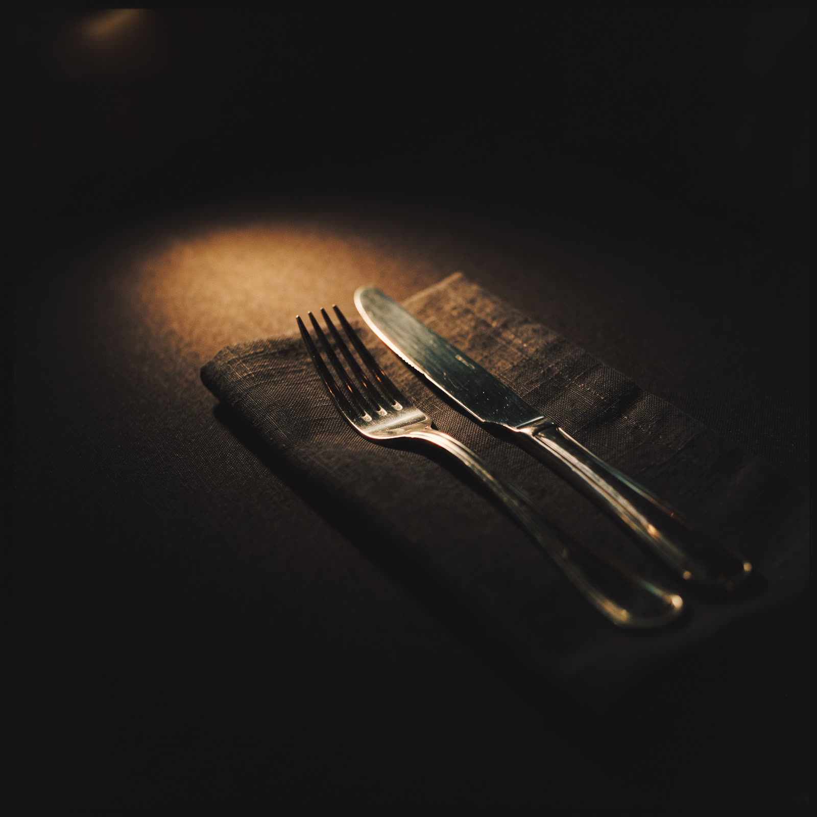 A silver fork and knife on a folded dark napkin, one hard side light
