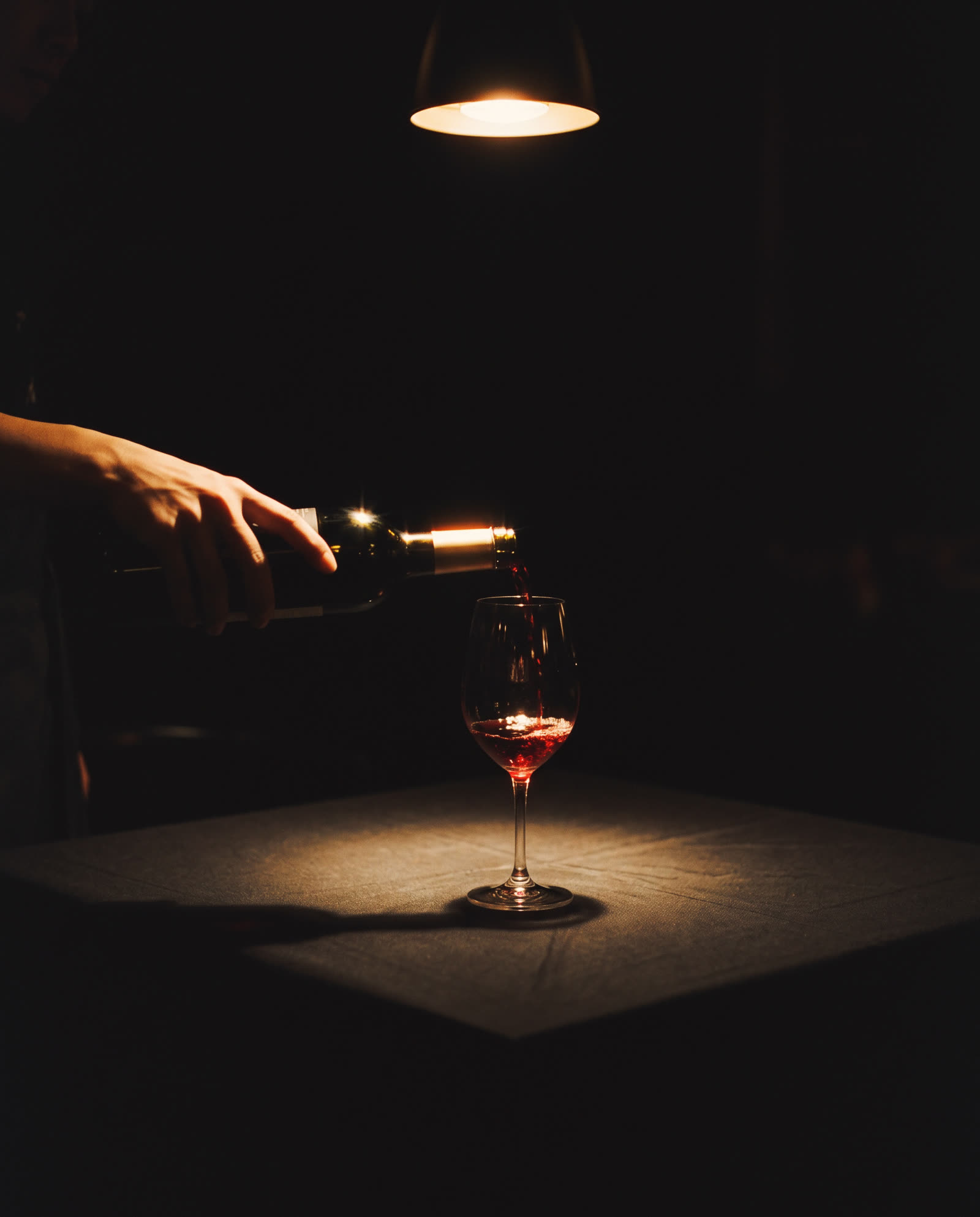 Red wine poured into one glass on a black tablecloth under a single lamp