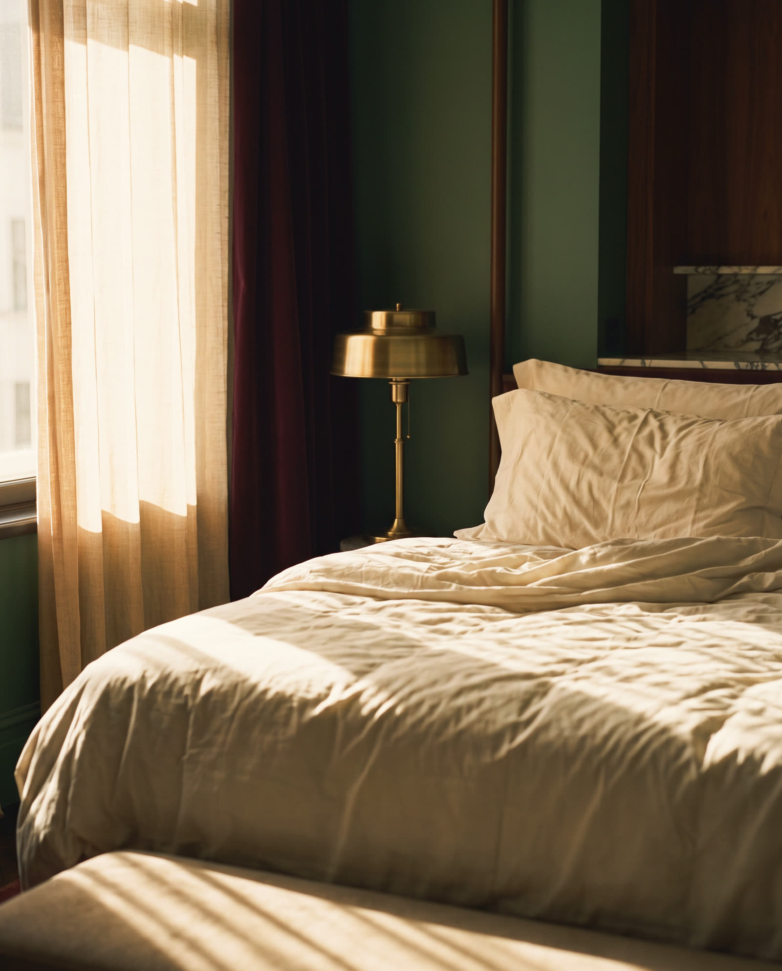 An unmade bed in cream linen, morning sun through the curtain