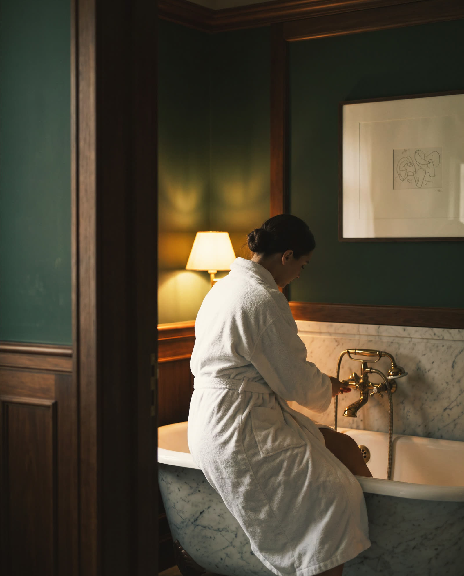 A woman in a white robe on the edge of a marble bathtub, seen from behind