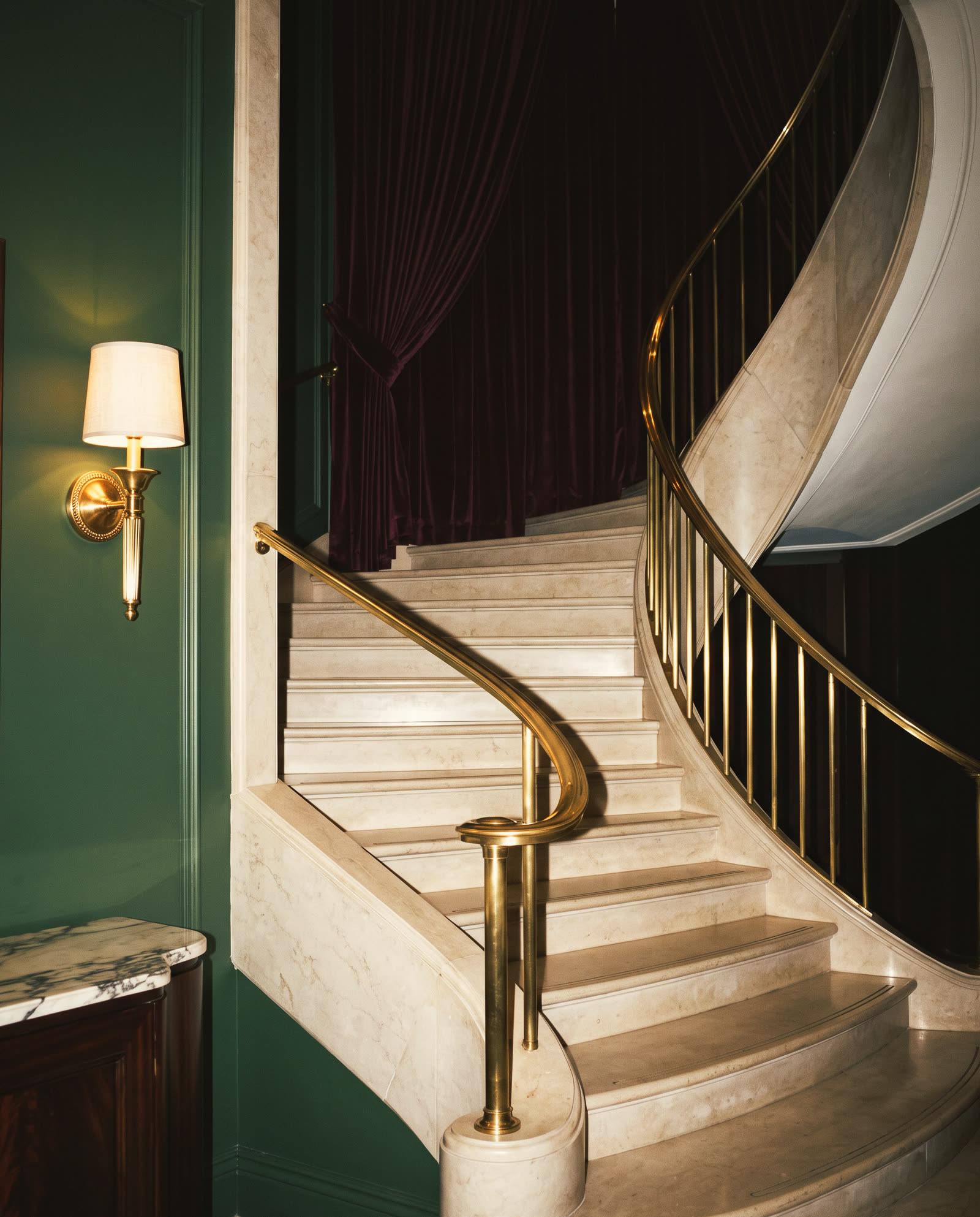 A curved stone staircase with a brass handrail and one wall lamp